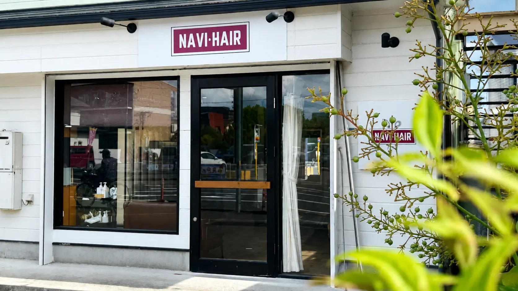 植物のグリーン越しに見える佐賀市赤松の美容室NAVI・HAIRの白い店舗外観